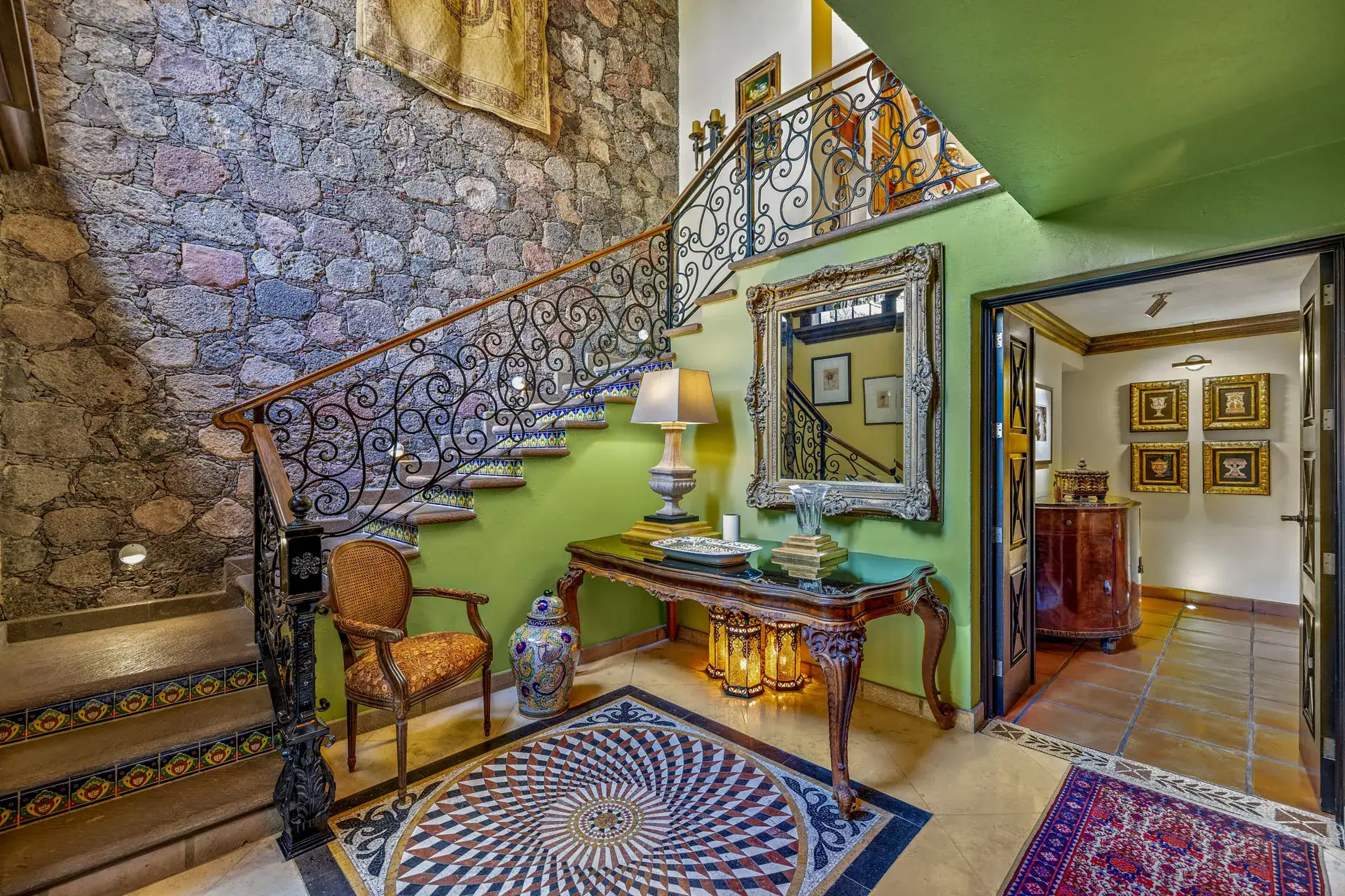Casa Flora, foyer with a wrought-iron staircase, a stone wall, a green wall and an ornate mirror over a console, San Miguel de Allende