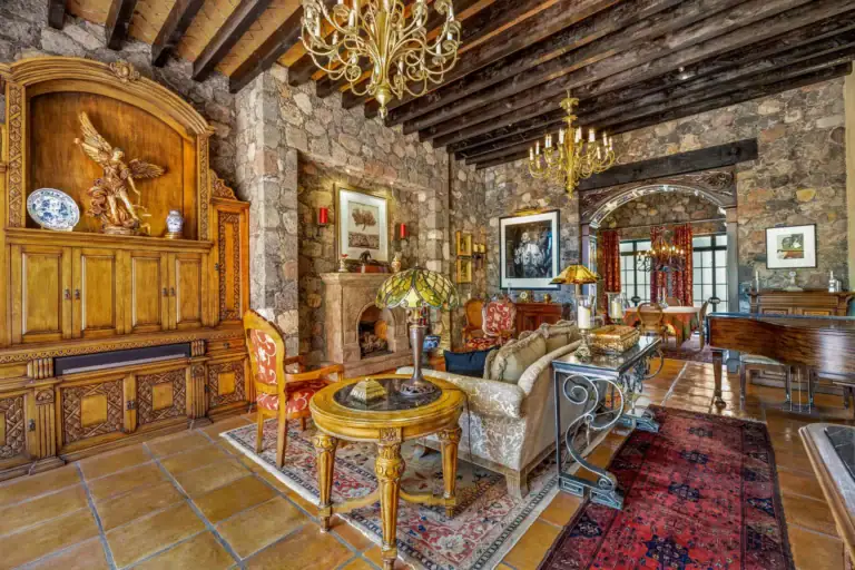 Casa Flora, great room with a stone wall, a carved wood cabinet, brass chandeliers and antique furniture under a beamed ceiling, San Miguel de Allende