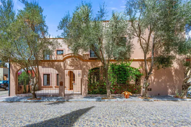 Casa Fuente del Sol, peach street wall with a wrought-iron gate and arched grille, olive trees and a cobblestone street, San Miguel de Allende