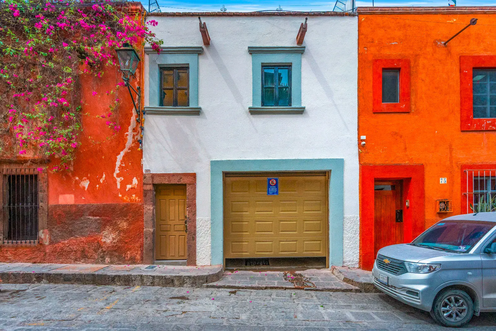 Casa Hidalgo, white and blue-trimmed two-story house with a tan garage door between an orange house and a red house with bougainvillea, San Miguel de Allende