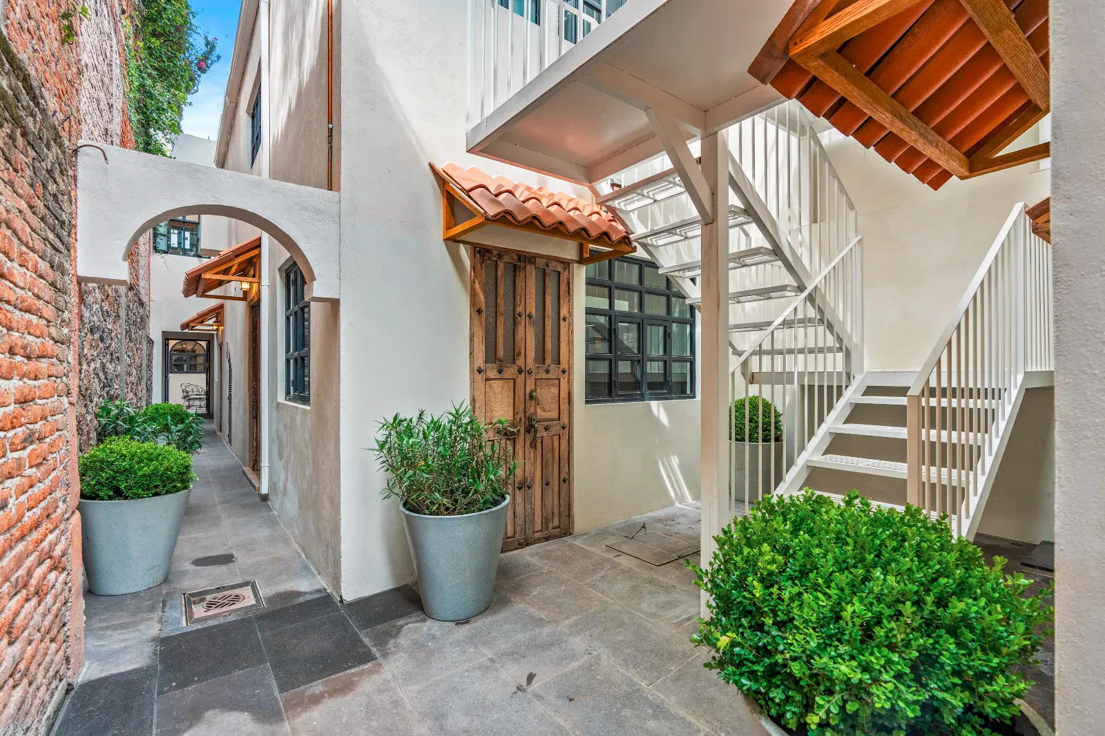 Casa Hidalgo, narrow courtyard passage with a carved wood door, a white metal staircase, potted shrubs and a tile roof canopy, San Miguel de Allende