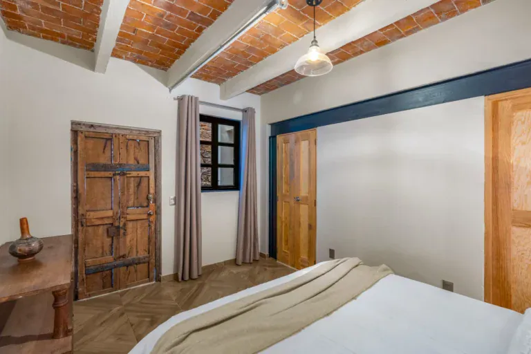Casa Hidalgo, bedroom with a carved wooden door, a white bed and a brick vaulted ceiling with white beams, San Miguel de Allende