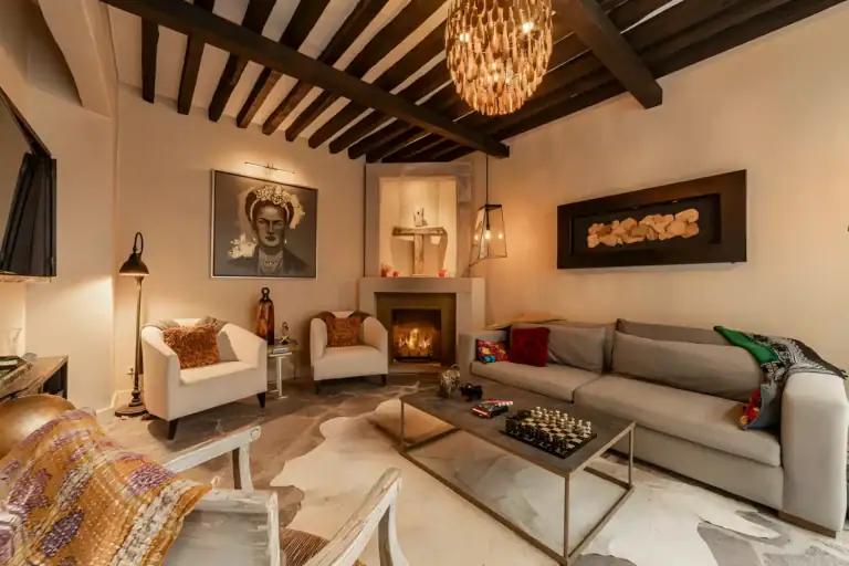 Casa Huerta, living room with a gray sofa, a corner fireplace, white armchairs, a dark beam ceiling and a shell chandelier, San Miguel de Allende