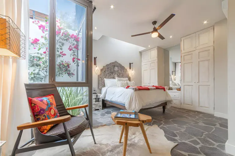 Casa Huerta, bedroom with a carved headboard, a ceiling fan, white wardrobes and a lounge chair by a window with bougainvillea, San Miguel de Allende