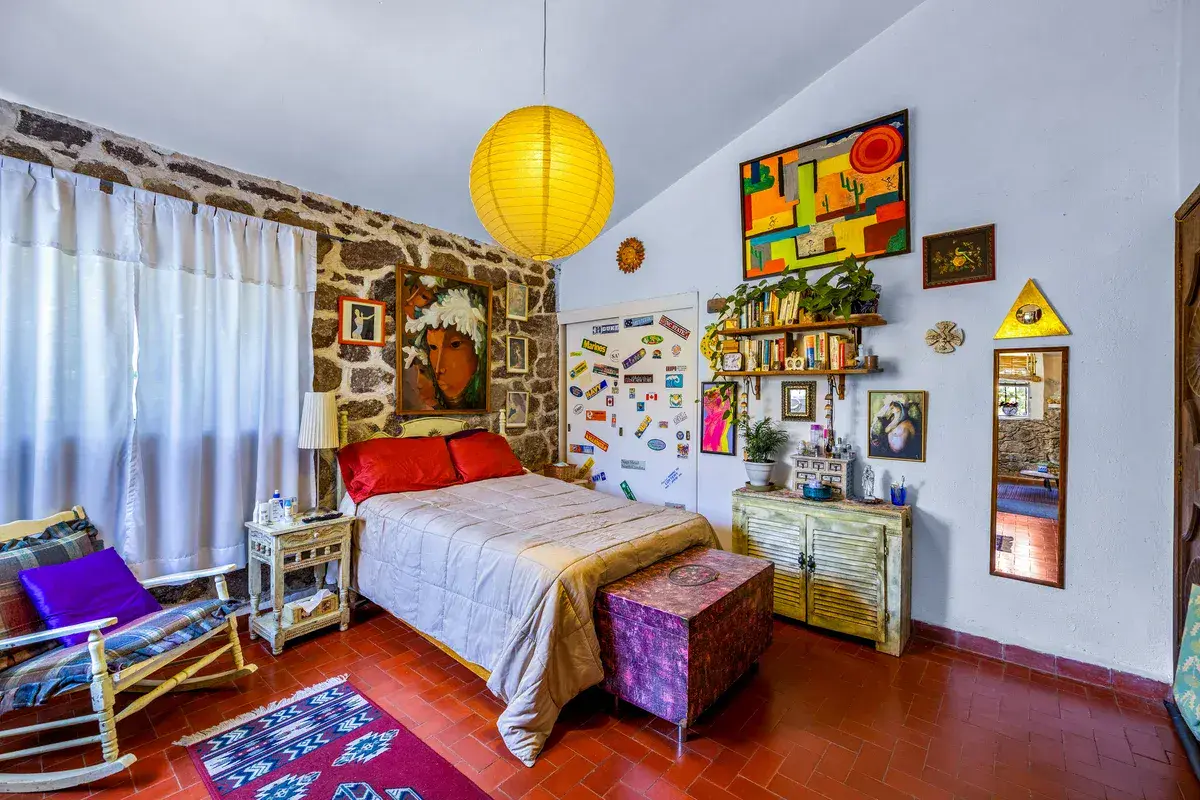 Casa La Chiquita, bedroom with a rocking chair, a bed with a red pillow, a stone wall and a colorful painting, San Miguel de Allende