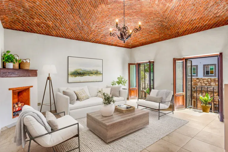 Casa Las Monjas, living room with a brick vaulted ceiling, a white sofa, armchairs, a wood coffee table and a corner fireplace (virtually staged), San Miguel de Allende