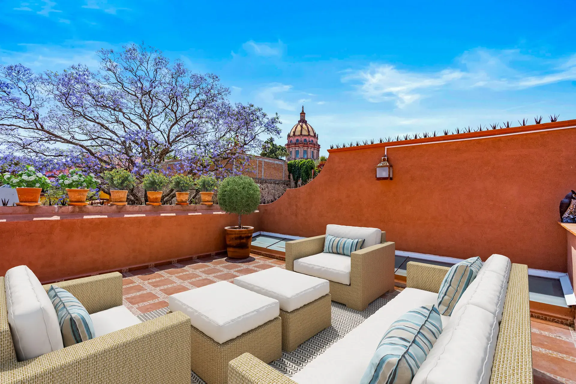 Casa Las Monjas, rooftop terrace with wicker sofas and ottomans, a terracotta wall and a view of a church dome and a flowering jacaranda tree (virtually staged), San Miguel de Allende