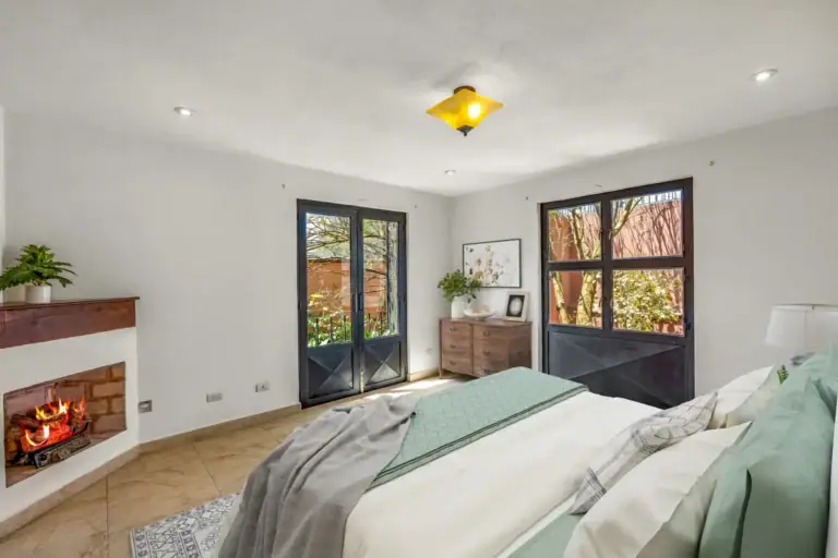 Casa Las Monjas, bedroom with a made bed with a sage green throw, a corner fireplace and French doors to a balcony (virtually staged), San Miguel de Allende