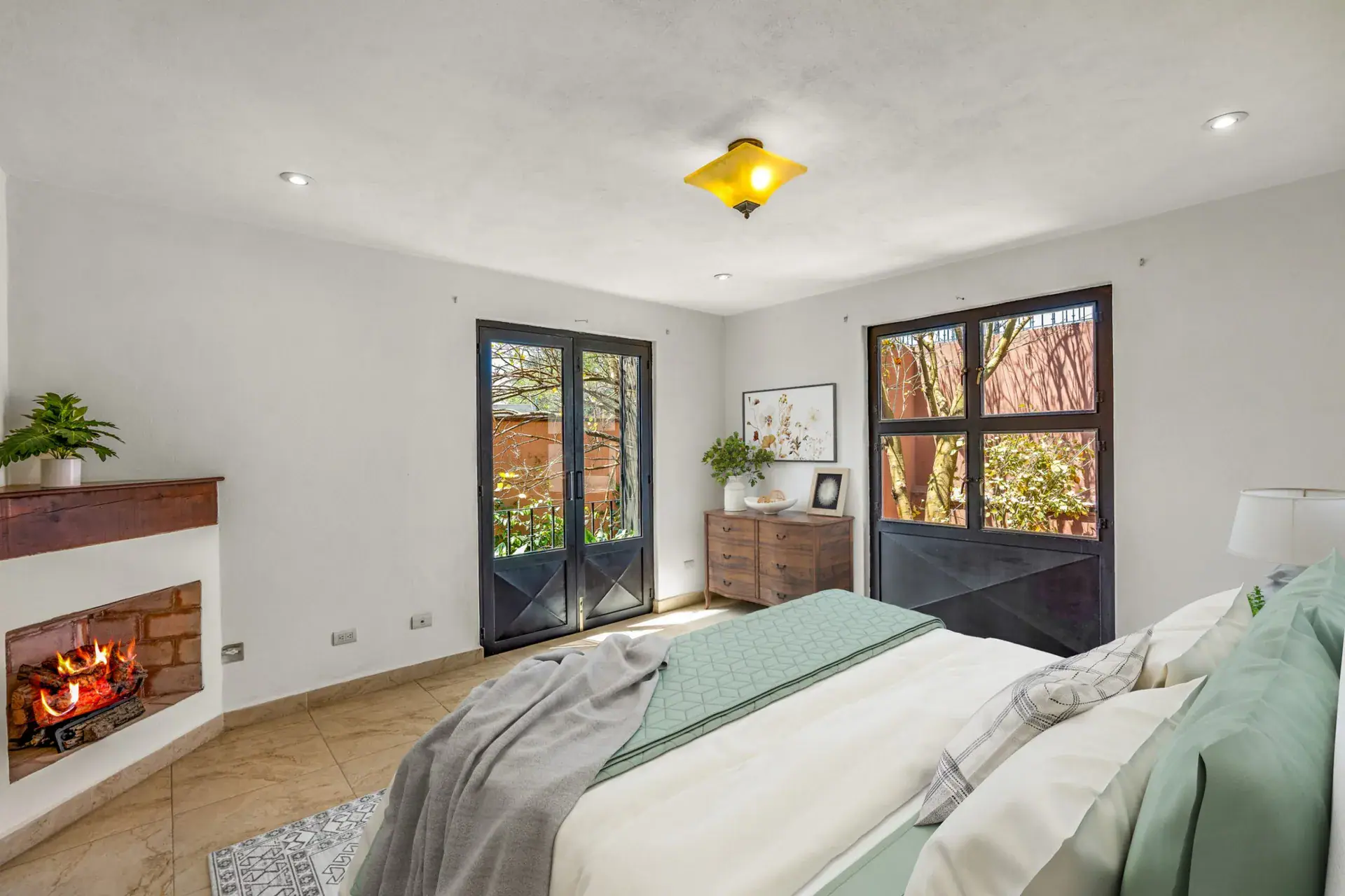 Casa Las Monjas, bedroom with a made bed with a sage green throw, a corner fireplace and French doors to a balcony (virtually staged), San Miguel de Allende