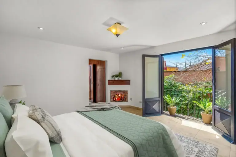 Casa Las Monjas, bedroom with a bed, a corner fireplace and open French doors to a leafy balcony (virtually staged), San Miguel de Allende