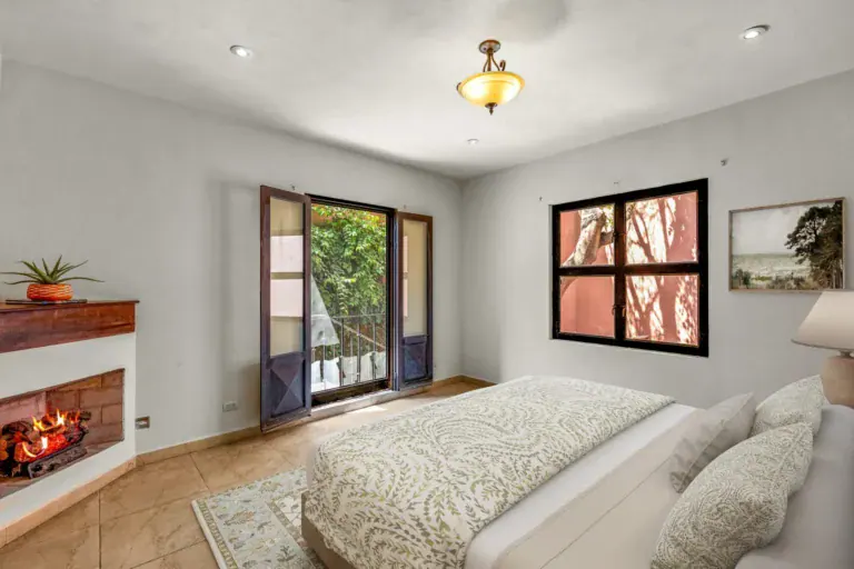 Casa Las Monjas, bedroom with a patterned bedspread, a corner fireplace and a window with a black frame (virtually staged), San Miguel de Allende