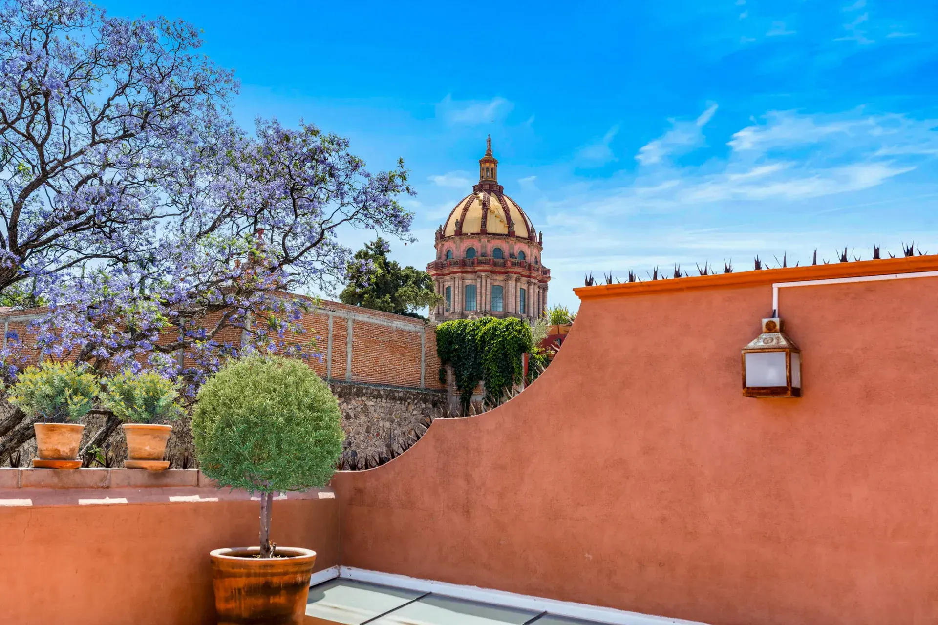 Casa Las Monjas, terrace with potted plants, a terracotta wall with a lantern and a church dome behind a jacaranda tree, San Miguel de Allende