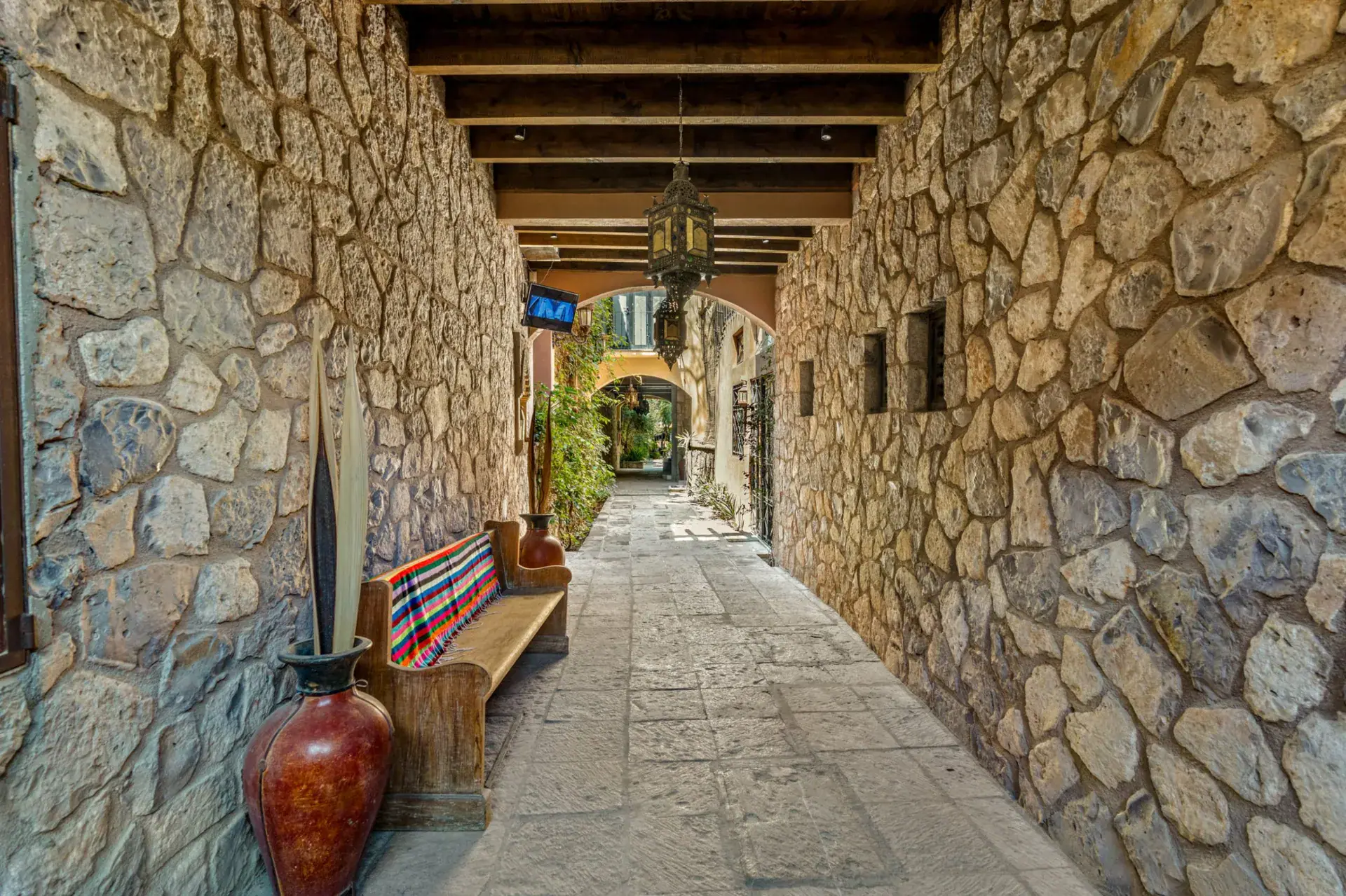Casa Las Monjas, covered stone passage with a wood beam ceiling, a bench with a striped blanket and a tall red vase, San Miguel de Allende