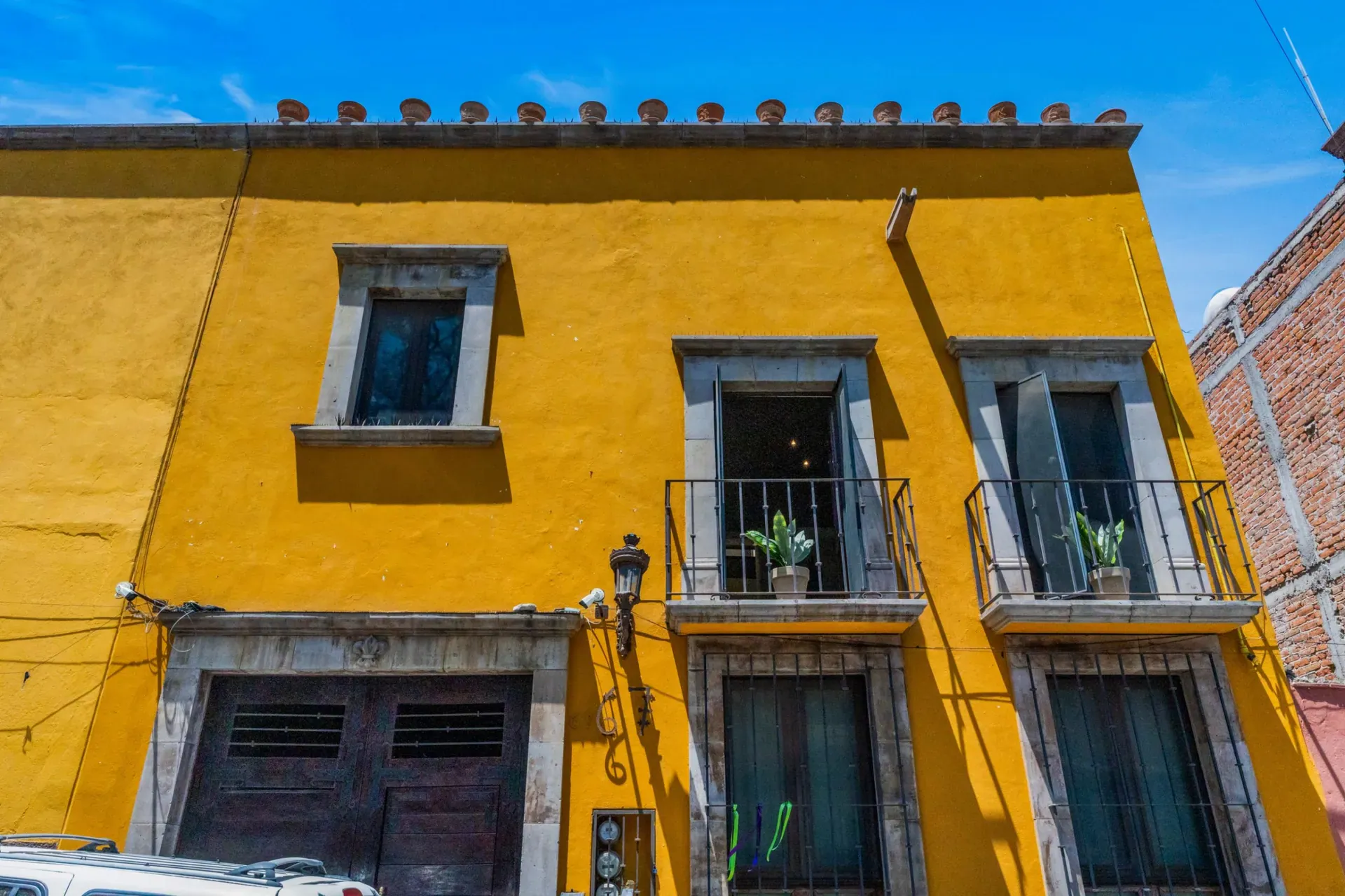 Casa Las Monjas, yellow facade with two balconies with plants, stone-framed windows and a tile cornice, San Miguel de Allende