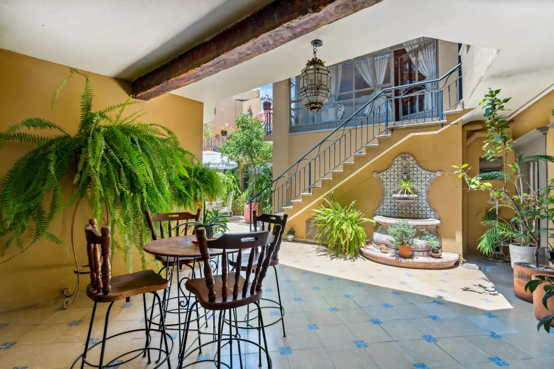 Casa Los Helechos, tiled courtyard with a bistro table, ferns, a tiled wall fountain and an iron staircase, San Miguel de Allende