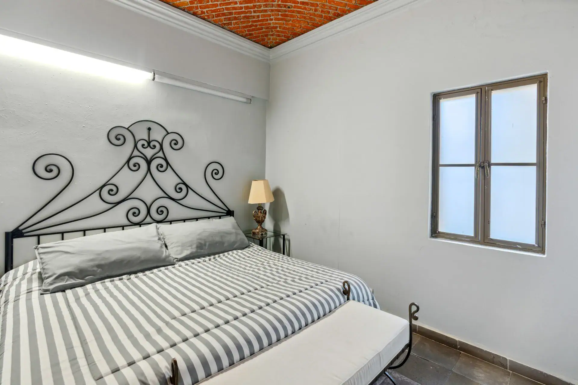 Casa Los Helechos, bedroom with a black iron headboard, striped bedding and a brick ceiling edge, San Miguel de Allende