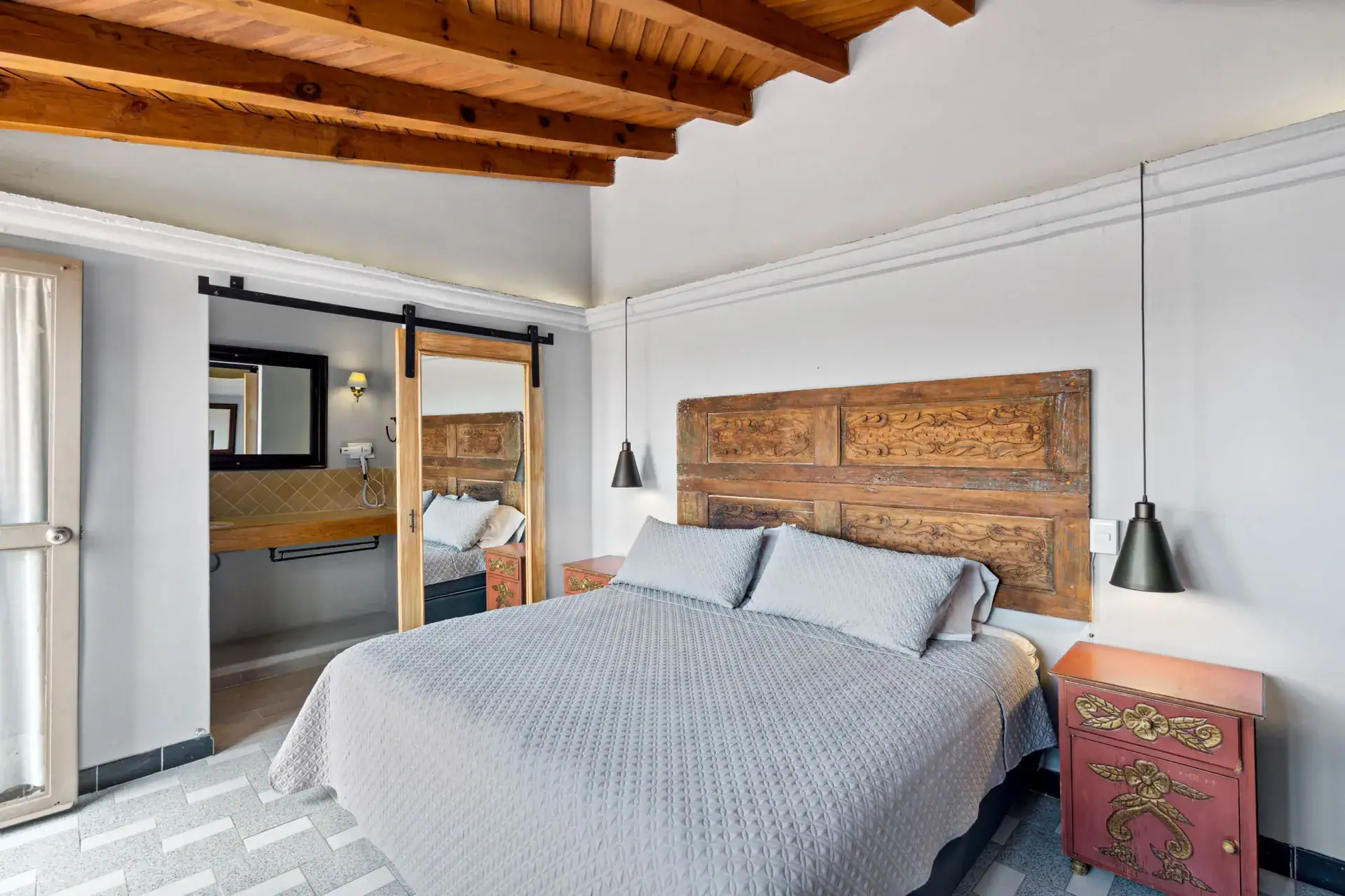 Casa Los Helechos, bedroom with a carved wood headboard, gray bedding and a barn-door bathroom, San Miguel de Allende