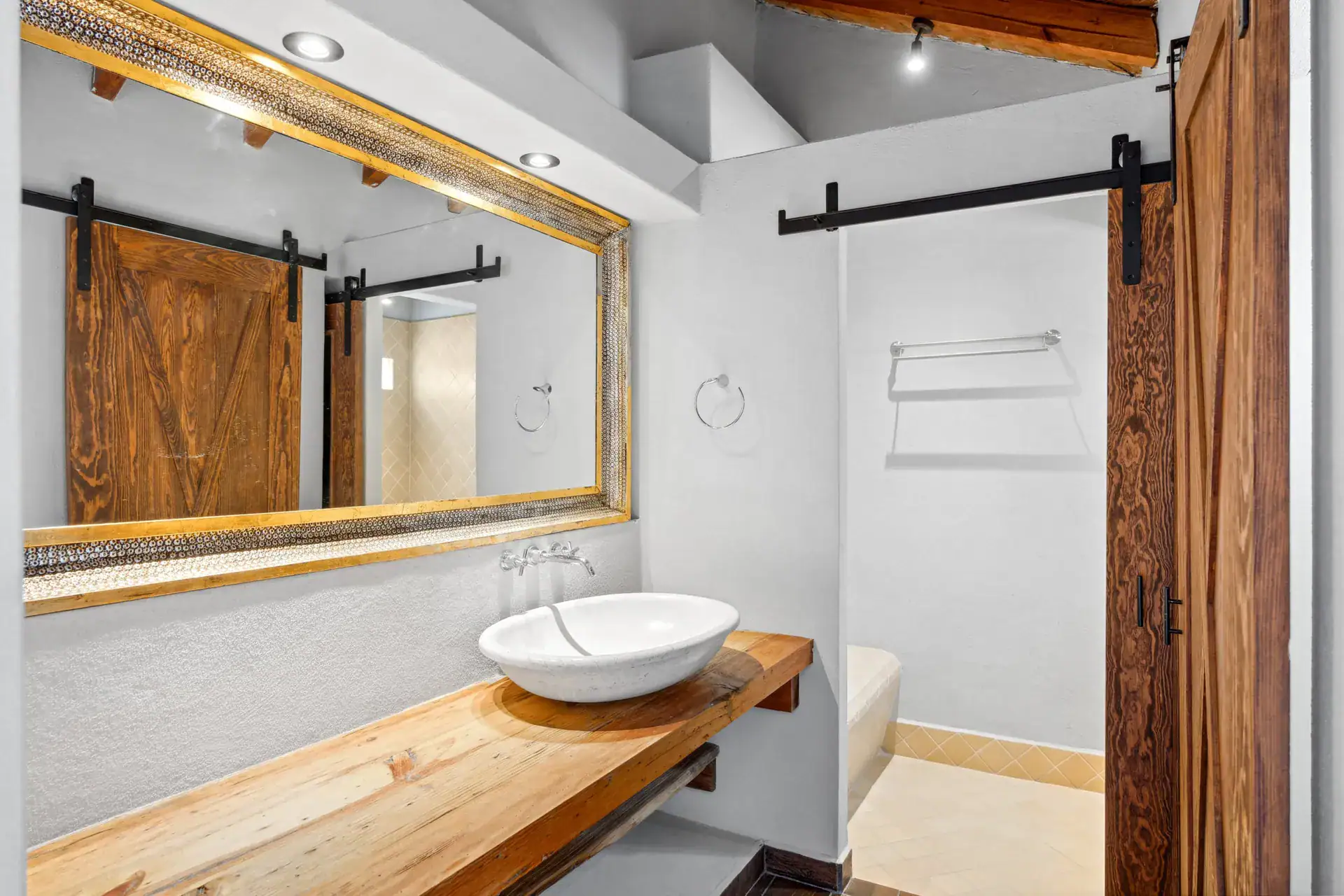 Casa Los Helechos, bathroom with a stone vessel sink on a wood counter, a gold-framed mirror and barn doors, San Miguel de Allende