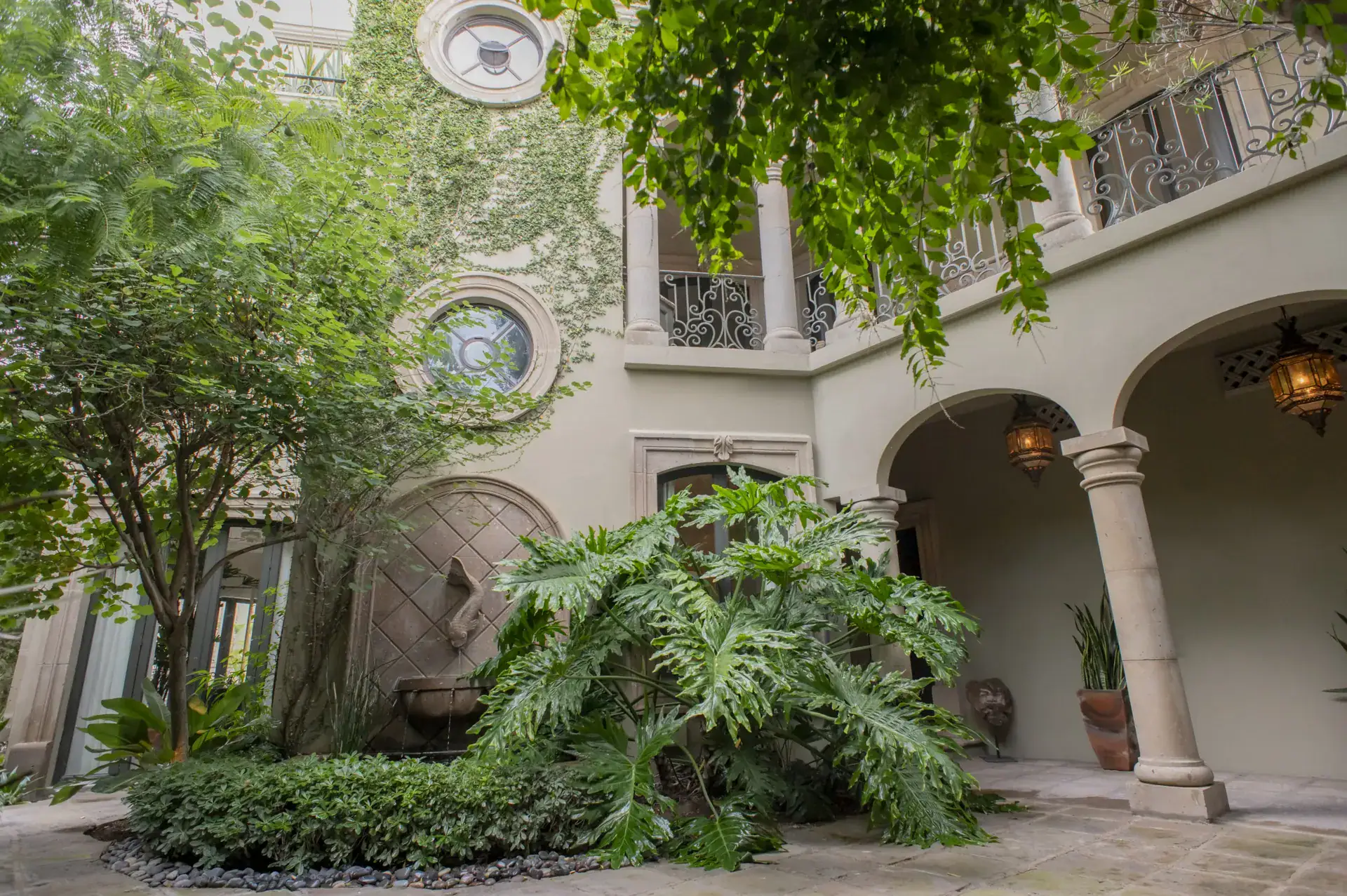 Casa Lyla, courtyard arcade with stone columns, a hanging lantern and lush foliage under a wrought-iron balcony, San Miguel de Allende