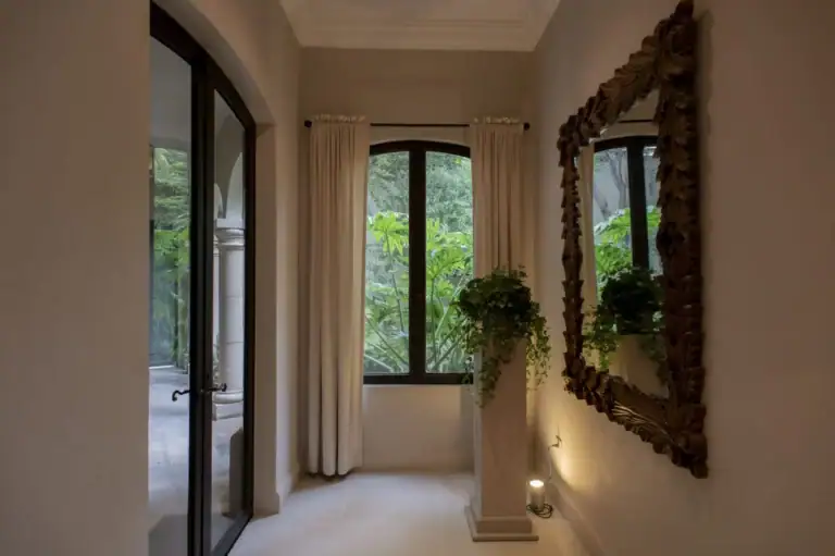 Casa Lyla, hallway with a carved wood mirror, a plant on a pedestal and arched windows to the garden, San Miguel de Allende