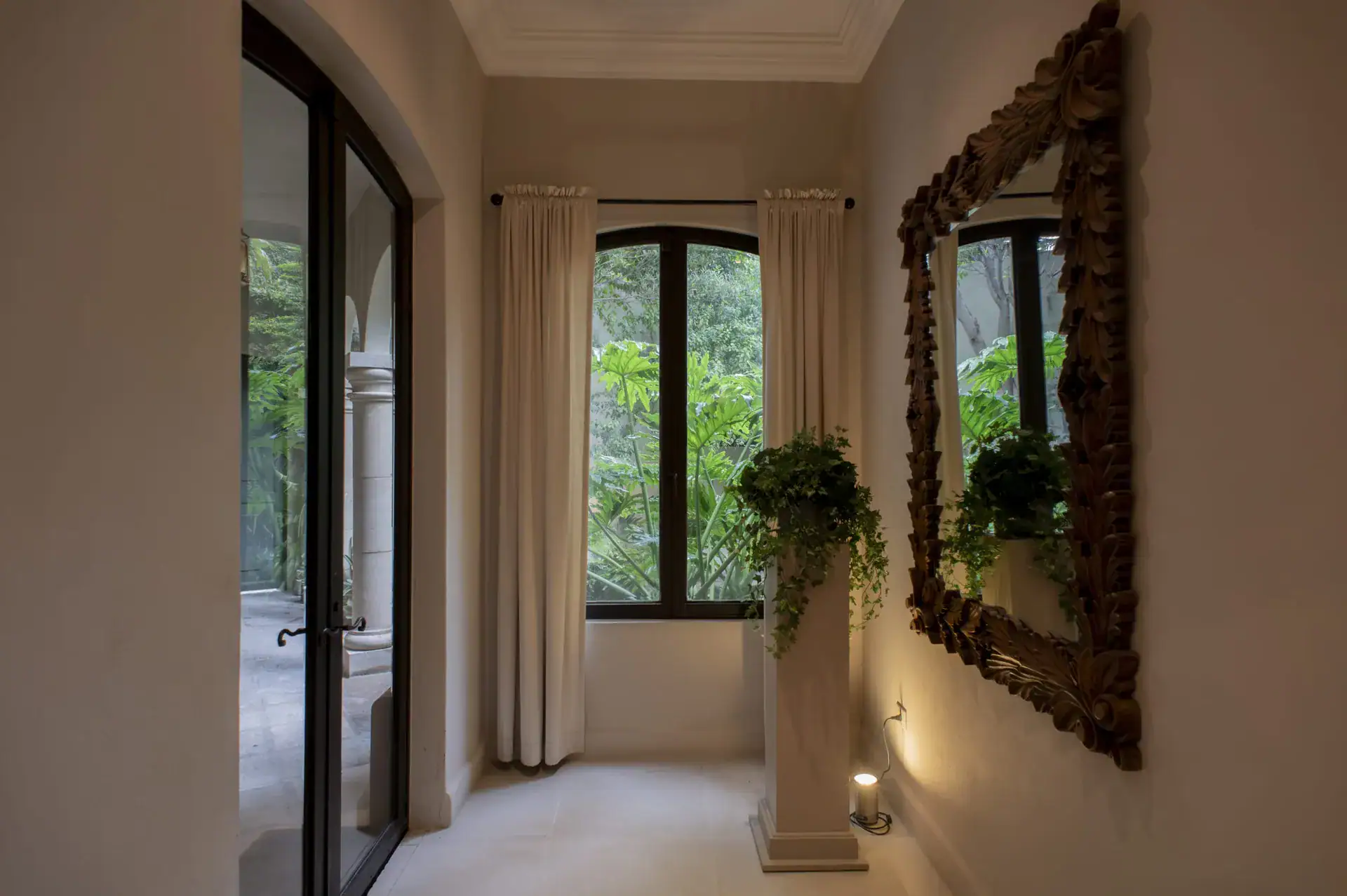Casa Lyla, hallway with a carved wood mirror, a plant on a pedestal and arched windows to the garden, San Miguel de Allende