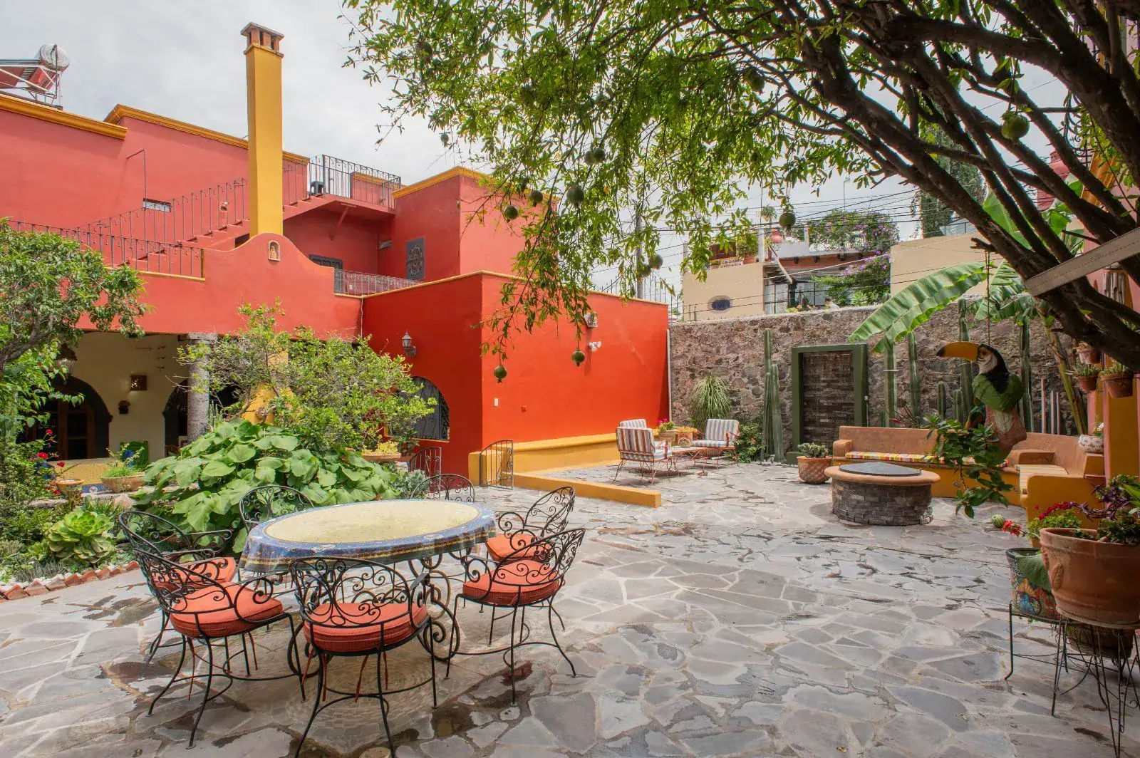 Casa Mexicana, courtyard with a wrought-iron table and chairs, a stone patio, tall trees and a red house, San Miguel de Allende