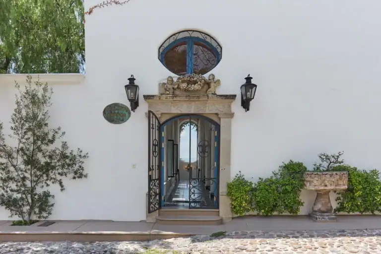 Casa Mi Besito, white entry wall with a stone-framed wrought-iron gate, an oval window and two lanterns on a cobblestone street, San Miguel de Allende