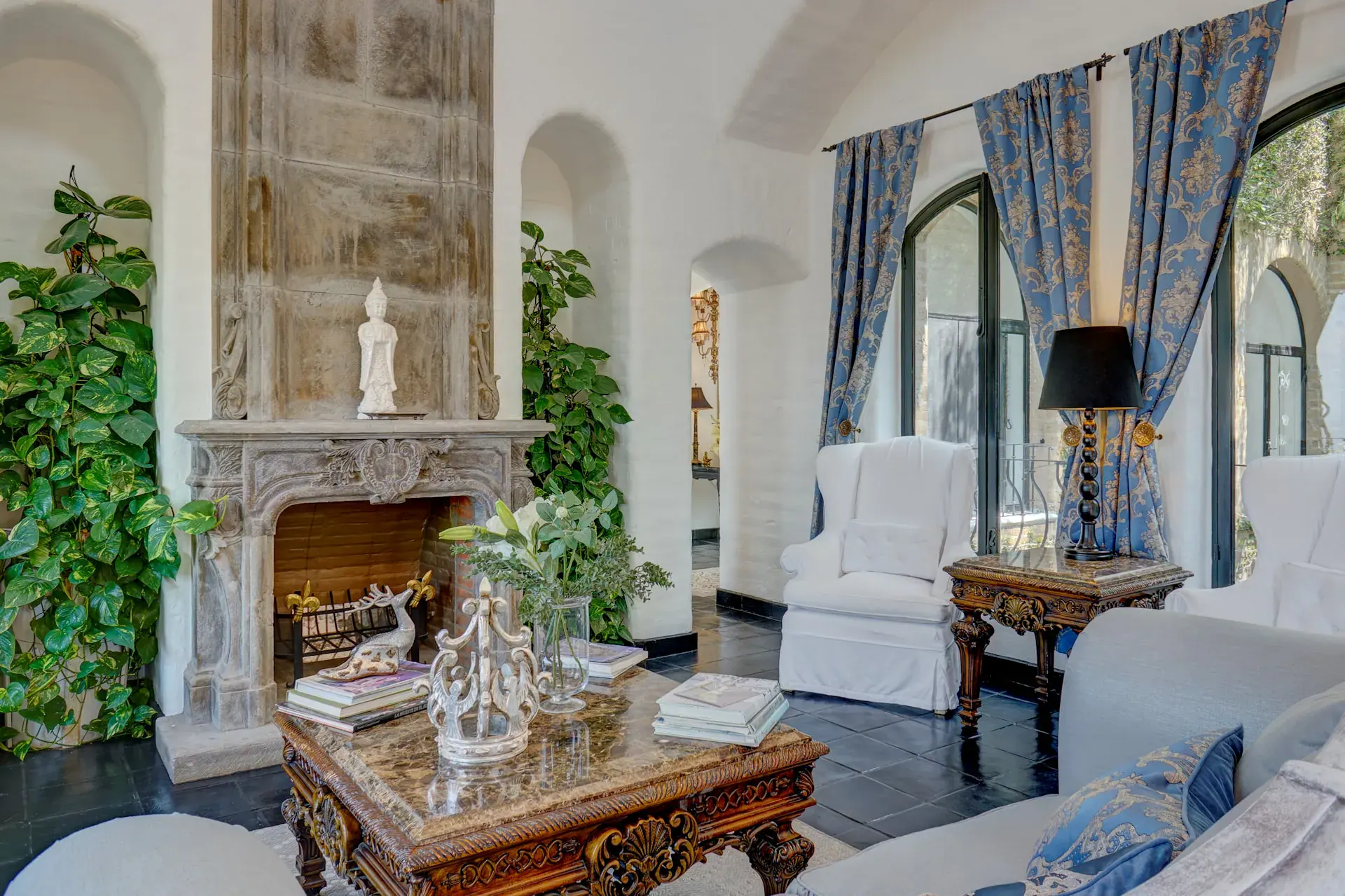 Casa Mi Besito, living room with a carved stone fireplace, white armchairs and arched windows with blue curtains, San Miguel de Allende