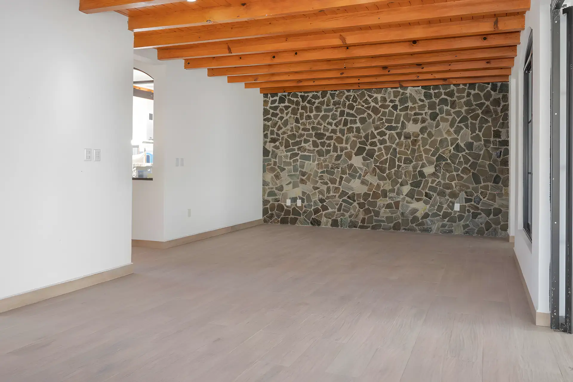 Casa Milagros, empty living room with a rough stone feature wall, a wood beam ceiling and a pale wood floor, San Miguel de Allende