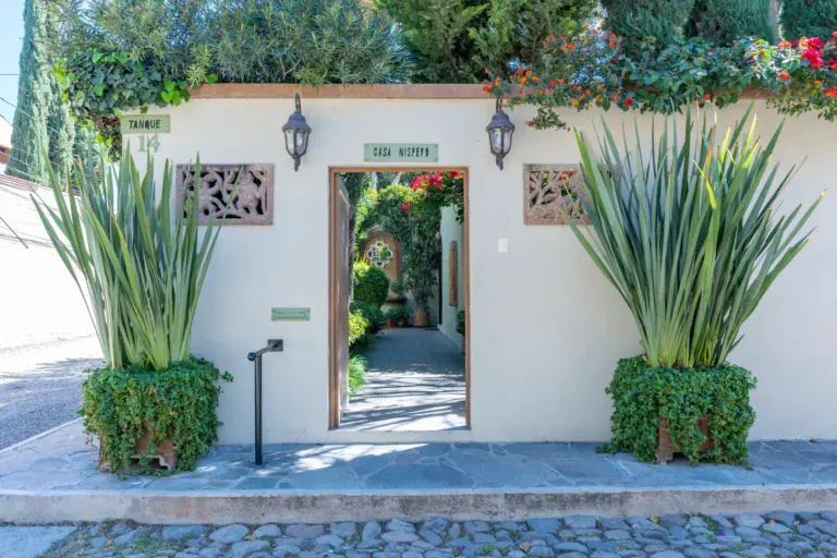 Casa Nispero, white garden wall with a doorway, a house name plaque and tall agaves in front on a cobblestone street, San Miguel de Allende