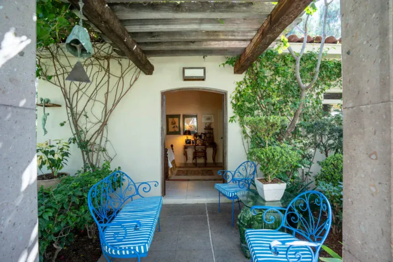Casa Nispero, covered walkway with blue iron benches, a bell and climbing vines leading to an arched doorway, San Miguel de Allende