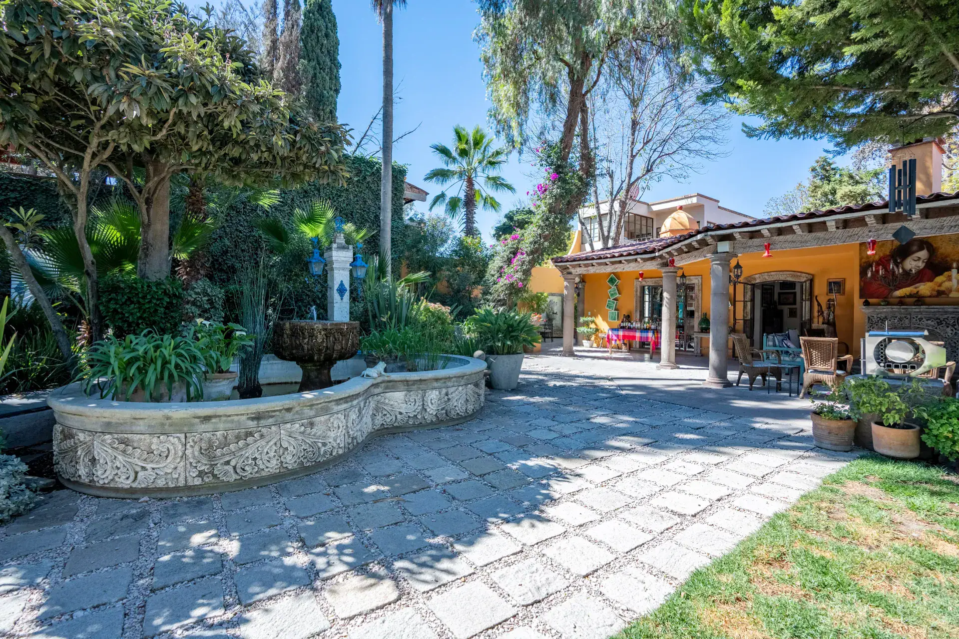 Casa Nispero, carved stone fountain basin in a paved garden with trees and the veranda behind, San Miguel de Allende
