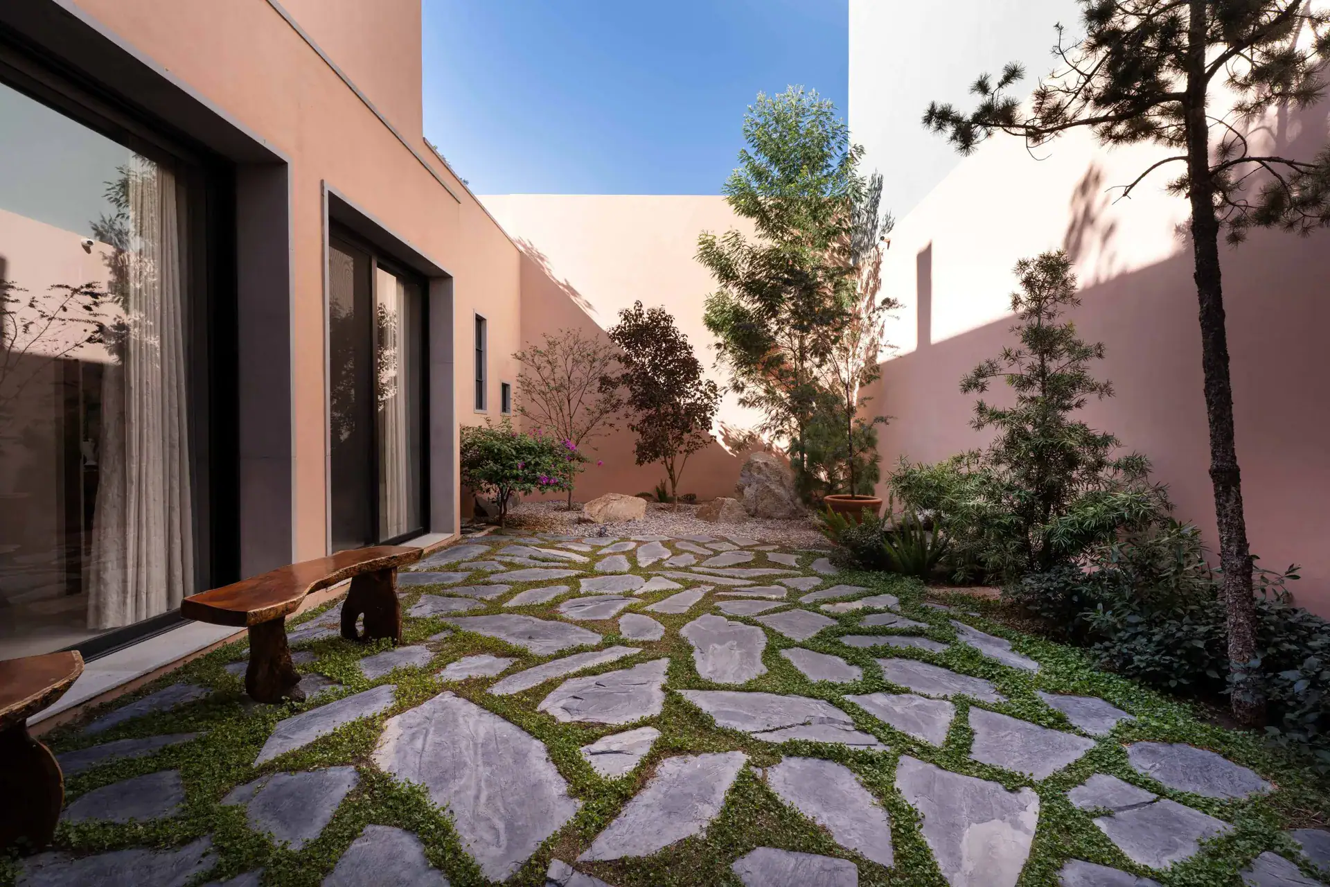 Casa Nobel, courtyard with a flagstone and grass pattern, a wood bench and pine trees beside a pink wall, San Miguel de Allende