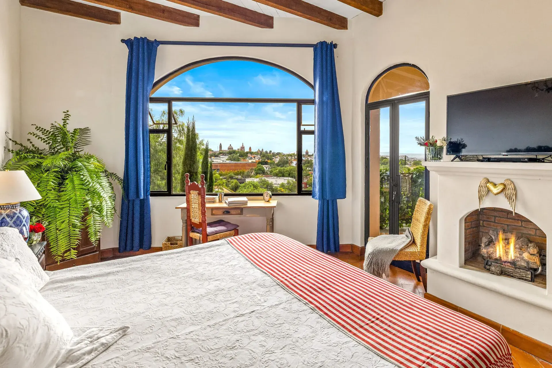 Casa Noche Magica, bedroom with a view through an arched window to treetops, a fireplace and a television, San Miguel de Allende