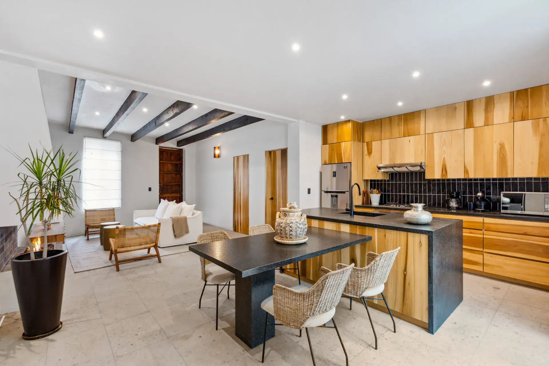 Casa Palma, open kitchen with natural wood cabinets, a black counter island, wicker chairs and a living area beyond, San Miguel de Allende
