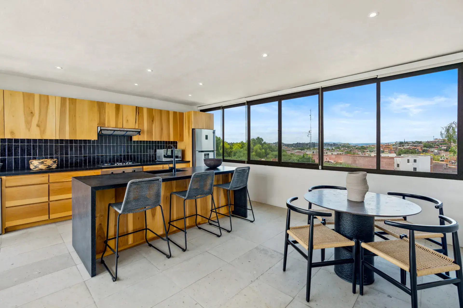 Casa Palma, upper kitchen with natural wood cabinets, a black island, wire stools and a round dining table by a wide window, San Miguel de Allende