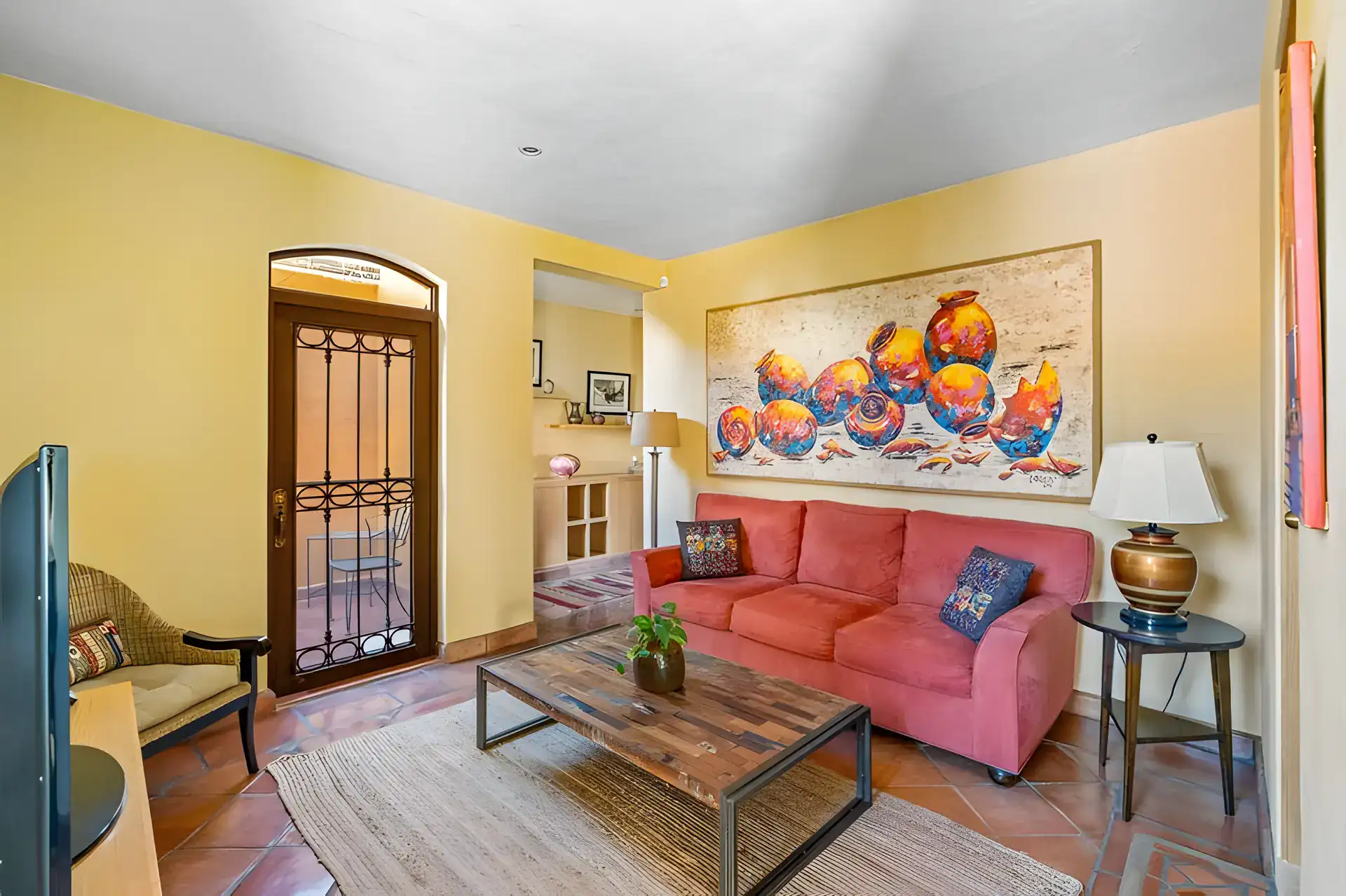 Casa Privada, den with a coral sofa, a large painting of fruit, a wood coffee table and an arched iron door, San Miguel de Allende