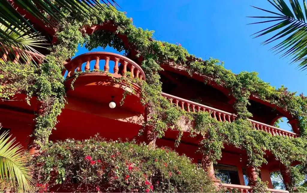 Casa Puesta del Sol, vine-covered balcony on the red facade under a blue sky, San Miguel de Allende