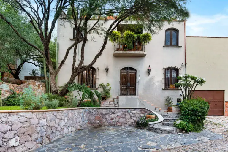 Casa Roka, white stucco house with arched windows, a balcony of plants, a wood door with stone steps and a mature tree, San Miguel de Allende