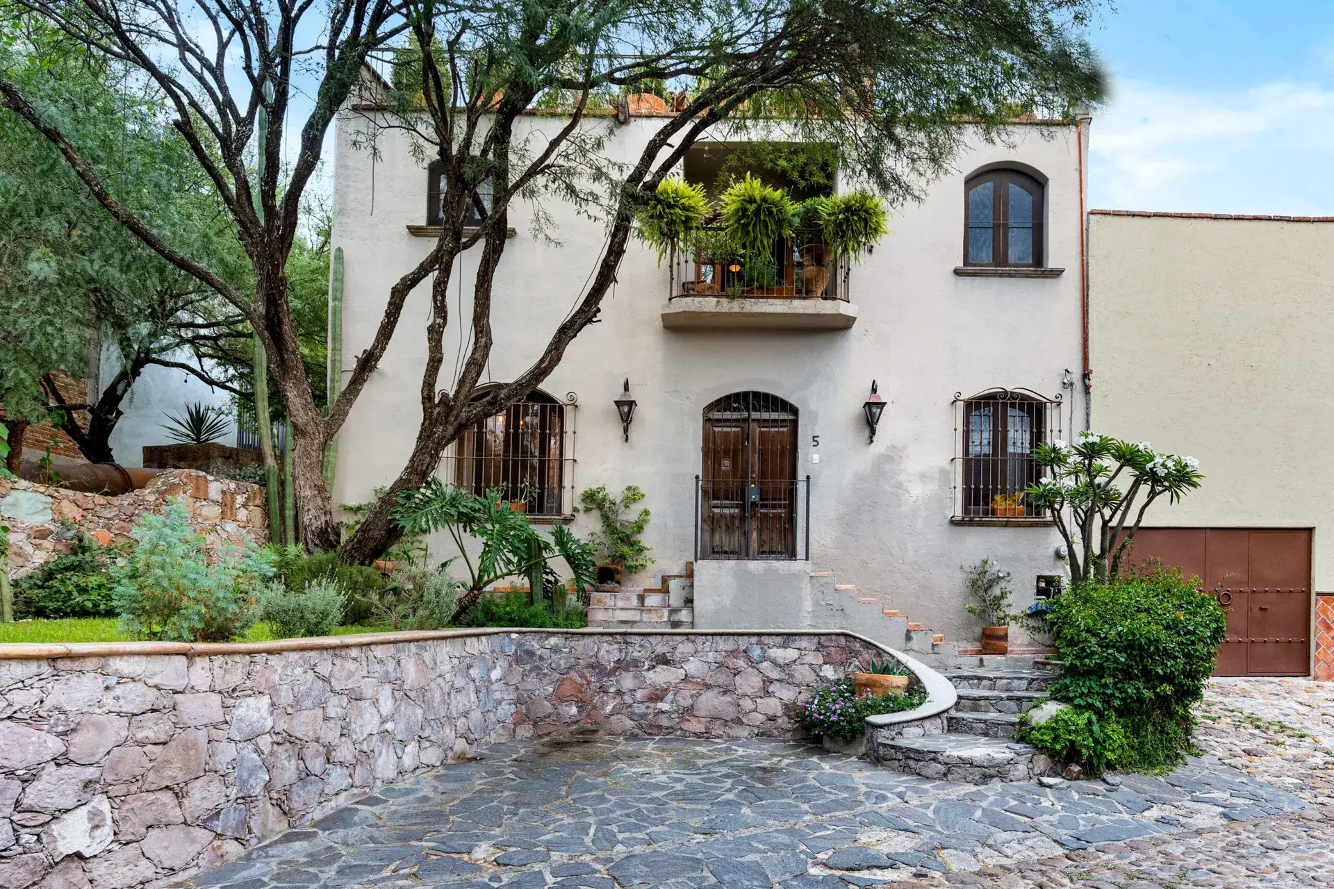 Casa Roka, white stucco house with arched windows, a balcony of plants, a wood door with stone steps and a mature tree, San Miguel de Allende