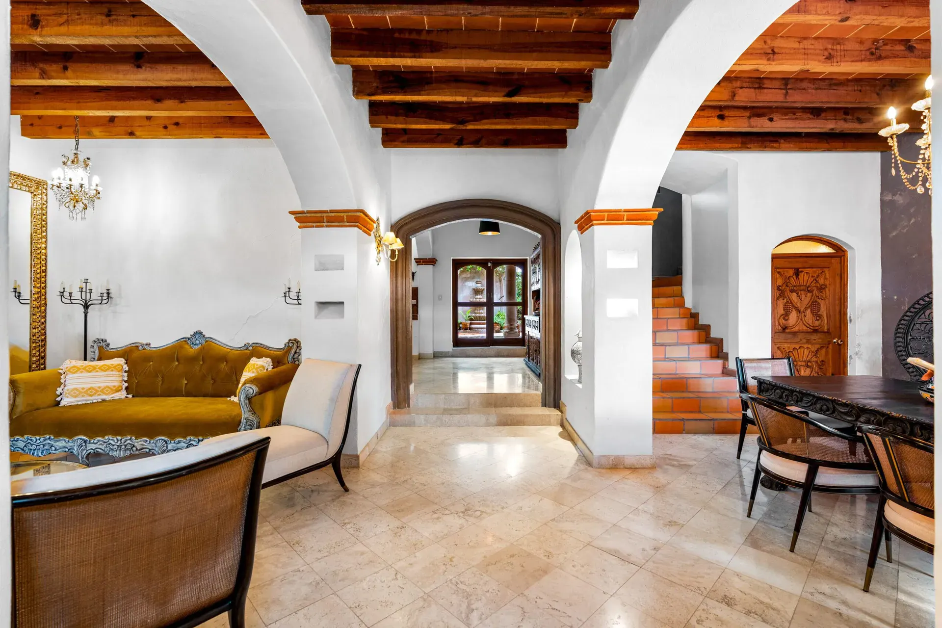 Casa Roka, living room with a gold velvet sofa, a carved wood hutch and a long dining table beyond arches with wood beams, San Miguel de Allende