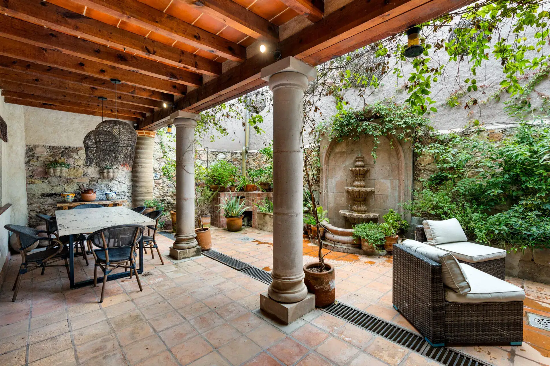Casa Roka, covered courtyard patio with stone columns, a dining table, a wicker sofa and a stone wall fountain, San Miguel de Allende