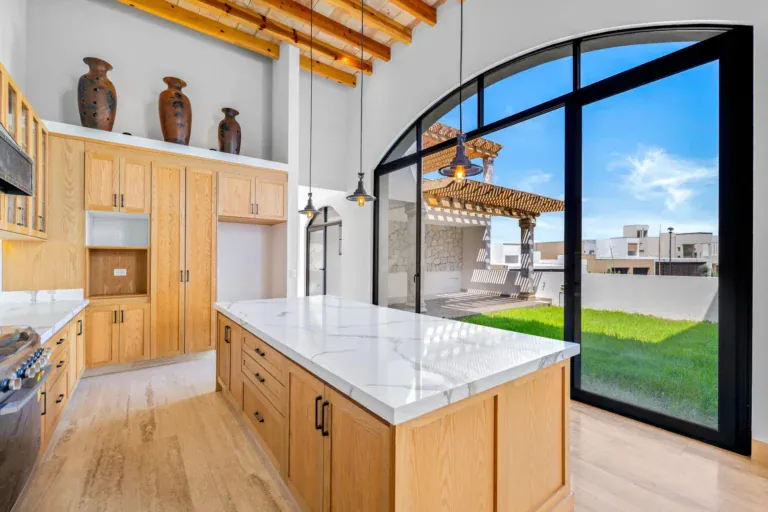 Casa Ronda, kitchen with a marble island, wood cabinets, a stainless range and an arched glass wall to a lawn and pergola, San Miguel de Allende