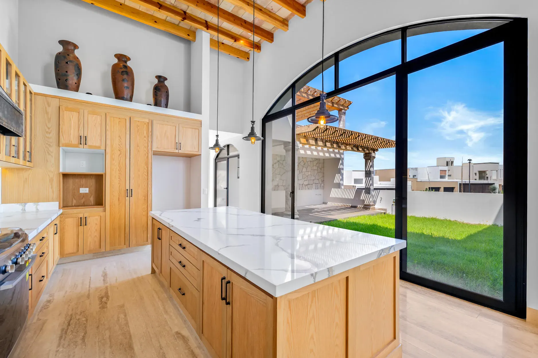 Casa Ronda, kitchen with a marble island, wood cabinets, a stainless range and an arched glass wall to a lawn and pergola, San Miguel de Allende