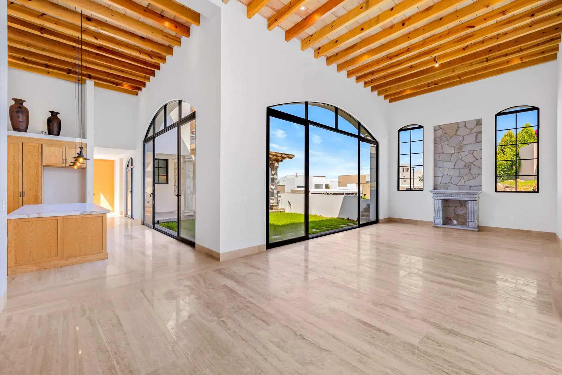 Casa Ronda, empty living area with a wood beam ceiling, a stone fireplace and an arched glass wall to the lawn, San Miguel de Allende