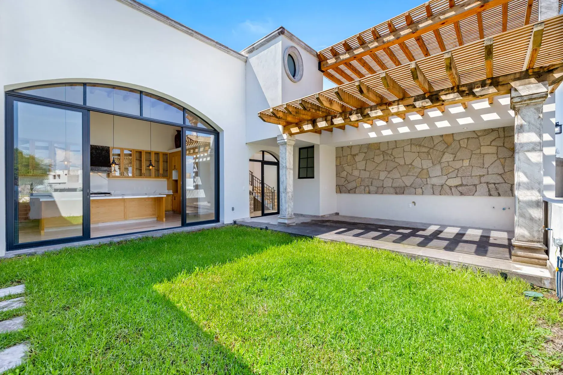 Casa Ronda, courtyard lawn with a covered outdoor kitchen under a wood pergola with a stone wall, and arched glass walls, San Miguel de Allende