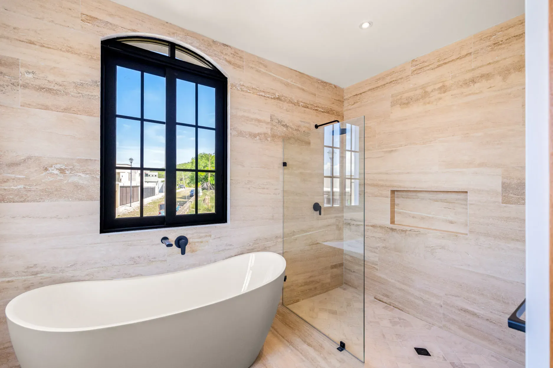 Casa Ronda, bathroom with a freestanding tub, a travertine wall and a black-framed arched window, San Miguel de Allende