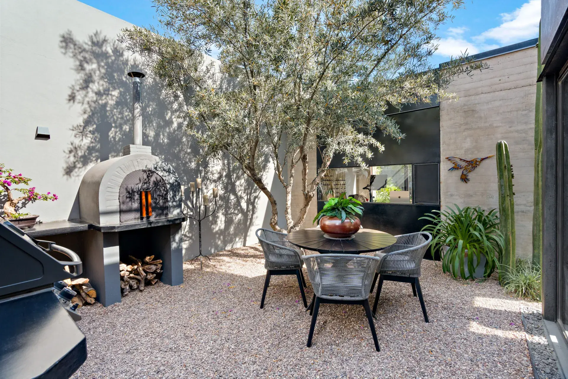 Casa Sanchez Renero, gravel patio with a wood-fired oven, a round table and a mature tree beside a gray wall, San Miguel de Allende