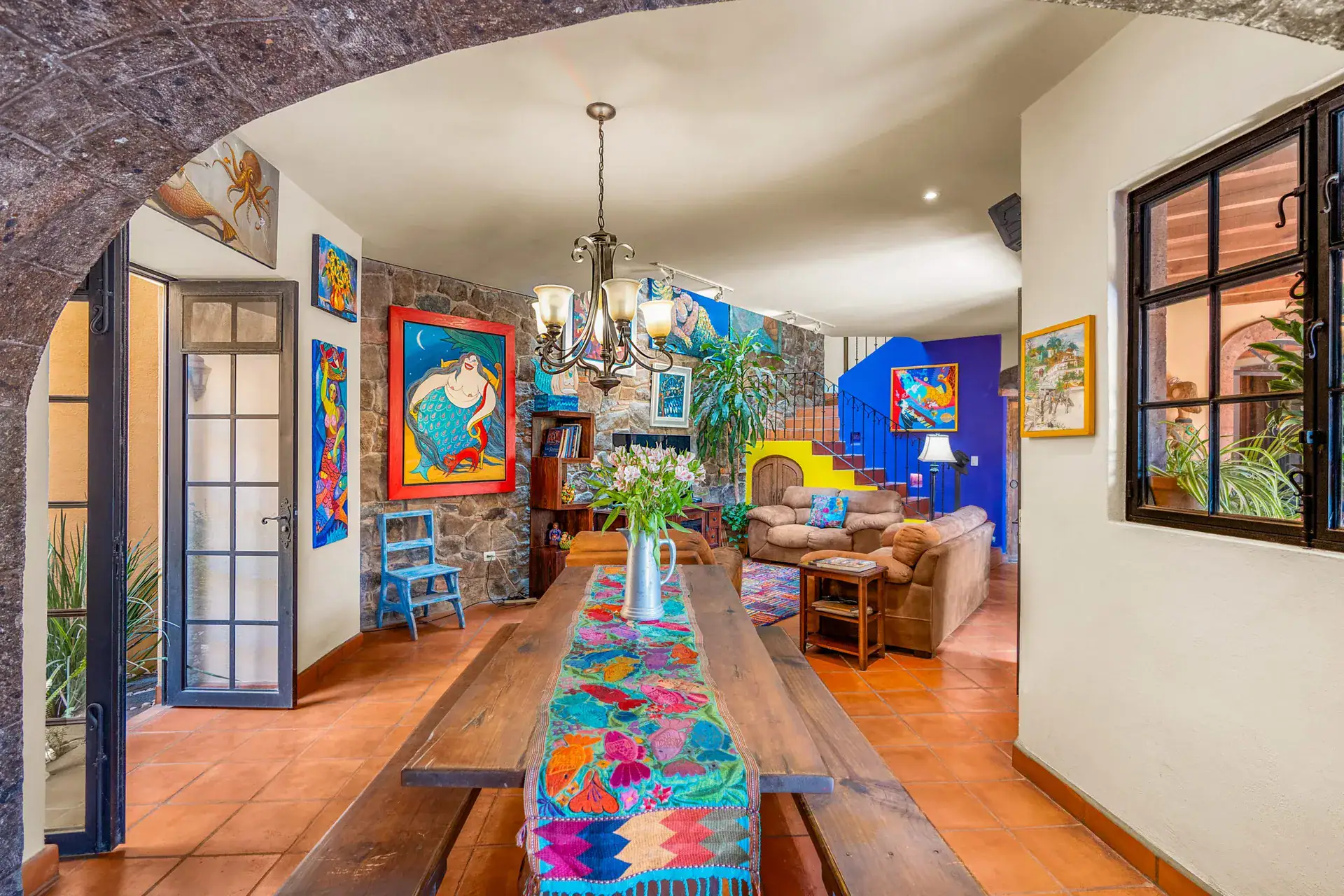 Casa Sirena, dining table with a colorful runner and benches, a stone wall, a blue art piece and a living room beyond, San Miguel de Allende