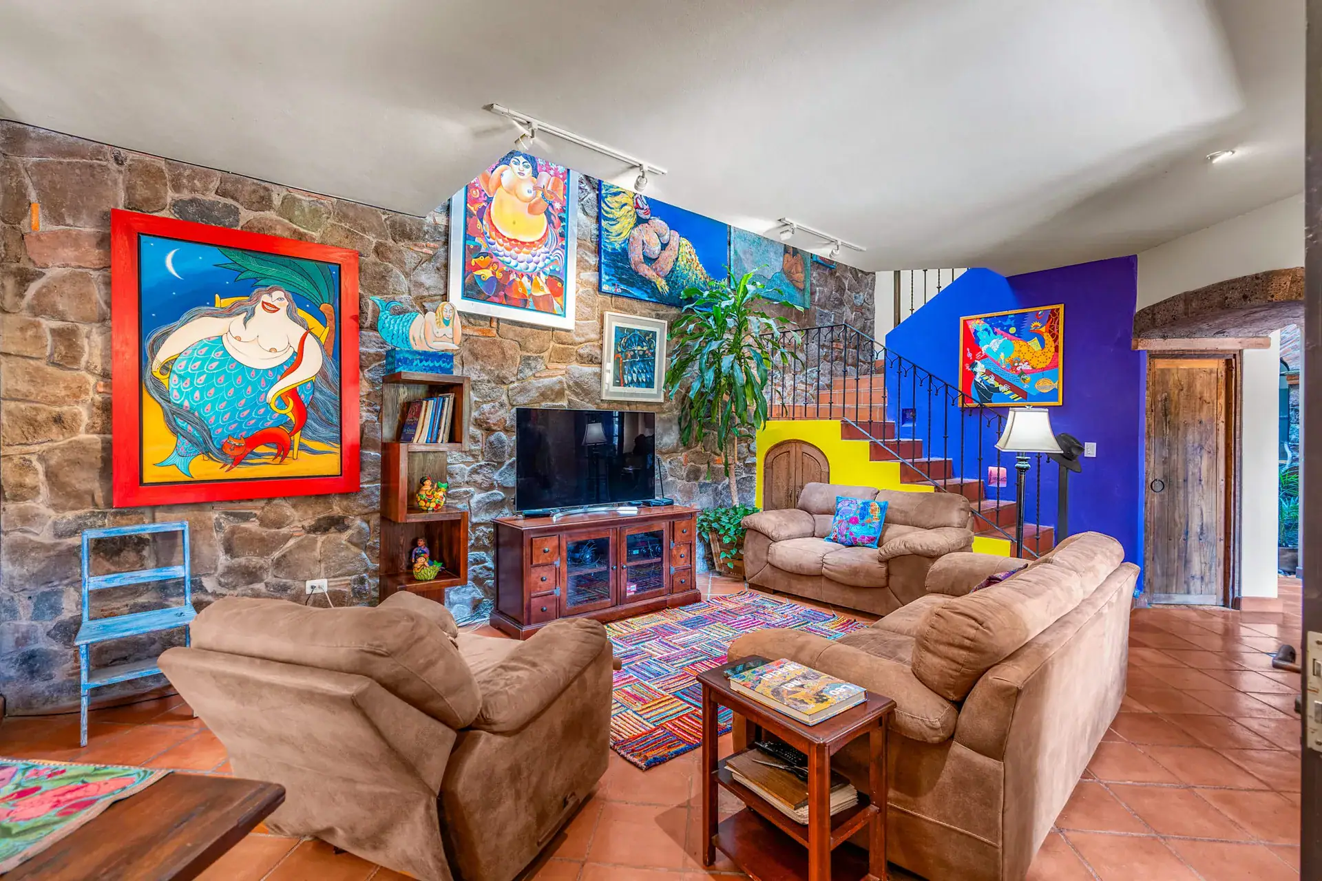 Casa Sirena, living room with a stone wall, a large painting of a woman, a television, brown sofas and a colorful rug, San Miguel de Allende